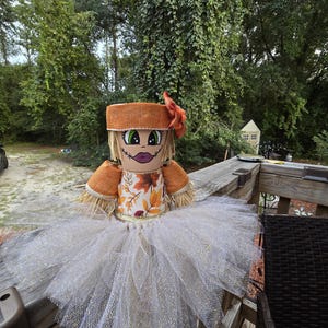 Könnte beinhalten: Eine dekorative Figur aus gestapelten Terrakottatöpfen mit einem bemalten Gesicht, grünen Augen und violetten Lippen. Sie trägt einen orangefarbenen Jutehut mit einer Blume, ein Oberteil mit herbstlichem Blattmuster und einen voluminösen, weiß glitzernden Tüllrock.