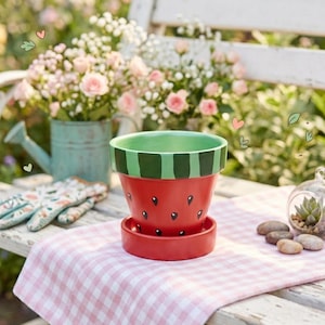 May include: A terracotta plant pot with a matching saucer, painted to resemble a watermelon. The main body is bright red with black teardrop-shaped seeds, while the rim features alternating light and dark green vertical stripes.