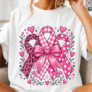 Op de afbeelding: Een wit t-shirt met een grafisch ontwerp van drie roze linten samengebonden met een centrale strik. De linten hebben luipaardprint, roze-witte ruitjes en roze stippen, omringd door kleine roze hartjes en sterretjes als decoratieve accenten.