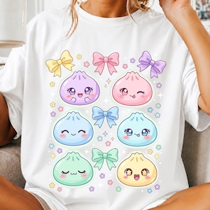 Può includere: Una t-shirt bianca con una stampa grafica di sei personaggi a forma di gnocco sorridenti con varie espressioni facciali. Il design è decorato con diversi fiocchi color pastello e piccole icone di fiori sparse tra gli gnocchi.