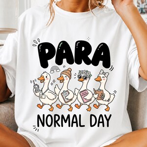 May include: A white t-shirt featuring a graphic of four cartoon ducks with the text PARA NORMAL DAY. The ducks are depicted with various accessories including a walkie-talkie, a beanie, sunglasses, a flower crown, and clipboards.