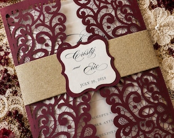 May include: A burgundy laser-cut wedding invitation with intricate floral patterns. The invitation is held together with a gold band and features the names "Cristy and Eric" and the date "July 20, 2019". The invitation is on a cream-colored background.