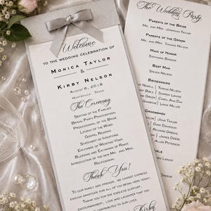 May include: Elegant wedding programs with a silver ribbon and bow, featuring the names Monica Taylor and Kirby Nelson, and the date August 6, 2018. The programs list the ceremony details and wedding party names. Soft pink roses and pearls accent the design.