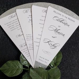 May include: A silver glitter wedding program fan with the text "Welcome to the Wedding Celebration of Monica and Kirby" and the date "August 6, 2018" and the location "Huntsville, Texas".