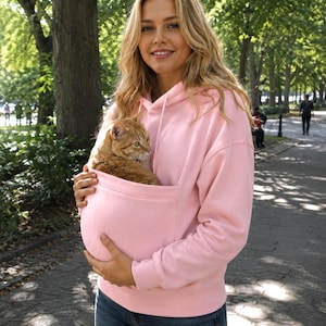 Op de afbeelding: Een lichtroze hoodie met een grote, geïntegreerde voorzak die is ontworpen om een klein dier in te dragen. De trui met lange mouwen heeft een capuchon met trekkoord en wordt gedragen met een donkerblauwe spijkerbroek, met een oranje kat erin.