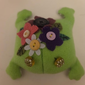 May include: A lime green felt pincushion shaped like a frog, decorated with fabric flowers in pink, white, and purple. The flowers are accented with various buttons, including a black button, a light purple button, and a yellow button.