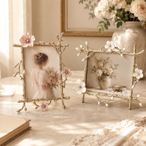 May include: Two gold-toned picture frames with a textured branch design. The frames are adorned with small, delicate white and pink floral accents. One frame is oriented vertically, and the other is oriented horizontally, both holding photos.