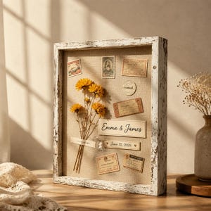 May include: A rectangular shadow box frame with a distressed white finish containing a tan linen backing. Inside are dried yellow flowers, vintage-style postage stamps, paper labels, and text reading Emma & James and June 15, 2026.
