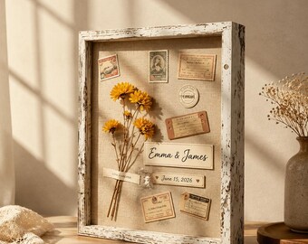 May include: A rectangular shadow box frame with a distressed white finish containing a tan linen backing. Inside are dried yellow flowers, vintage-style postage stamps, paper labels, and text reading Emma & James and June 15, 2026.