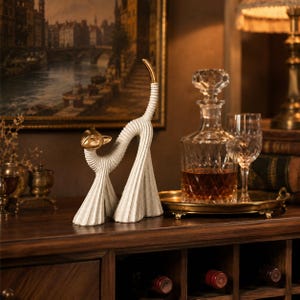 Peut inclure: Une figurine de chat décorative avec un corps blanc cassé texturé à stries verticales, une tête dorée lisse et une queue à pointe dorée. La sculpture adopte une pose arquée avec des pattes larges et évasées, alliant finitions métalliques et mates.