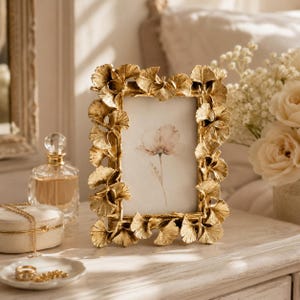 May include: A rectangular picture frame with a metallic gold finish, featuring a decorative border composed of sculpted ginkgo leaves. The frame holds a white insert displaying a single, delicate watercolour illustration of a pale pink flower.