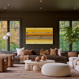 Op de afbeelding: Een rechthoekig abstract schilderij met horizontale strepen in tinten geel, oranje en grijs, opgehangen aan een donkere muur boven een bruine bank. Het kunstwerk toont gelaagde kleurbanden binnen een dunne omlijsting.