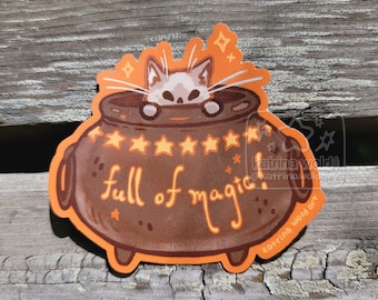 May include: A sticker featuring a brown cauldron with the words "full of magic" in orange script. A white cat peeks over the rim. Orange stars and sparkles decorate the design, with an orange outline.