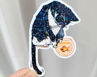 May include: A die-cut sticker featuring a black and white cat with a dark blue, star-patterned coat looking down at a small, round orange fishbowl containing a single orange goldfish. The text Katrina Wold Art is printed on the sticker.
