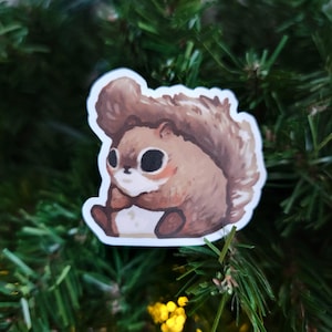 May include: A sticker featuring a cartoon squirrel with brown fur, large black eyes, and a white outline. The squirrel is sitting with its paws clasped, giving it a cute appearance. The sticker is set against a background of green pine needles.