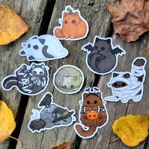 May include: A selection of Halloween cat stickers. The stickers depict cats in costumes such as a pumpkin, ghost, bat, skeleton, and mummy. The stickers are in various colours, including orange, black, white, and brown.