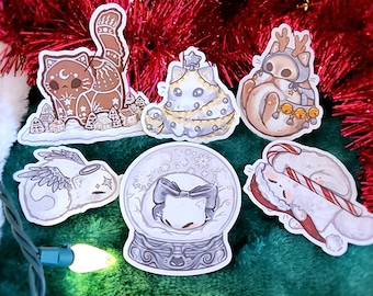 May include: A collection of whimsical cat stickers with a Christmas theme. The stickers feature cats in various festive designs, including a cat in a snow globe, a cat dressed as a Christmas tree, and a cat with reindeer antlers. The background is a green surface with red tinsel.