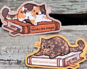 May include: Two cat stickers. One is a calico cat on a book with the text "books are magic". The other is a brown cat on a book with the text "read banned books". Both stickers have an orange border.