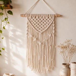 May include: A cream-colored macrame wall hanging featuring a triangular woven pattern suspended from a horizontal wooden dowel. The design includes intricate knots and long, loose fringe strands that hang vertically from the bottom edge.