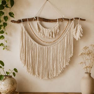 May include: A wall hanging featuring a natural wooden branch suspended by a cord. It includes cream-colored macrame fringe, a strand of light wooden beads, a smaller macrame crescent section, and a single macrame feather element attached to the branch.