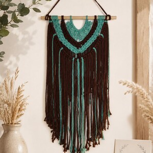 May include: A wall hanging made of dark brown and teal yarn featuring intricate macrame knots suspended from a horizontal wooden dowel with a yarn loop for hanging. The design includes layered V-shaped patterns and long, loose fringe.