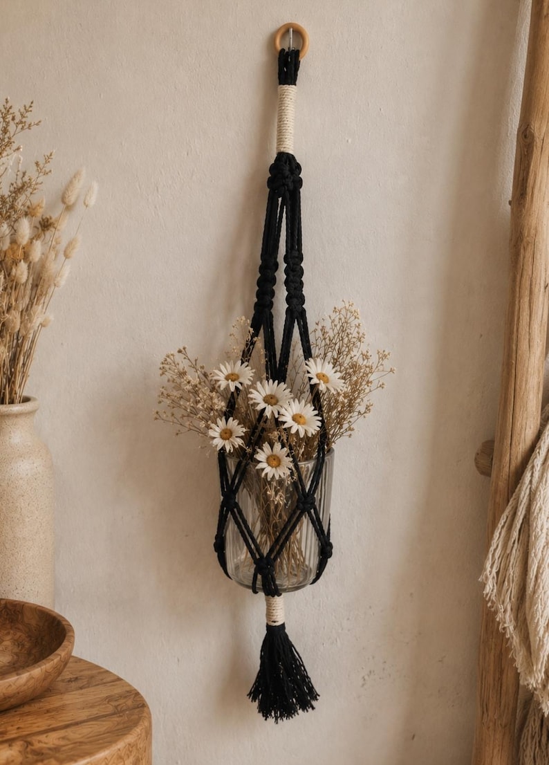 May include: A black macrame plant hanger featuring a knotted cord design with a cream-colored wrapped top and bottom tassel. The hanger holds a clear, vertically ribbed glass vase filled with white daisy flowers and dried botanical stems.