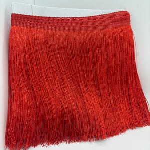 May include: A vibrant red fringe trim featuring a woven horizontal header band at the top. The trim consists of long, thin, straight vertical threads that hang down from the header, creating a dense and uniform texture across the entire piece.