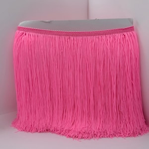 May include: A vibrant neon pink fringe trim featuring a dense row of long, thin, hanging threads attached to a woven header band. The material is uniform in color and texture, displaying a straight, vertical orientation across the entire length.