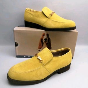 May include: A pair of bright yellow suede loafers featuring a gold-toned metal bit across the vamp and a dark brown sole. The shoes are displayed with a cardboard box that features a graphic of a Basset Hound and the partial text HUS.