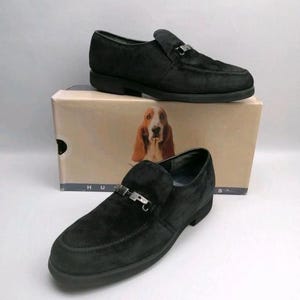 May include: A pair of black suede loafers featuring a silver-toned metal bit across the vamp. The shoes are positioned in front of a tan cardboard box that displays a graphic of a basset hound and the partial text "H U P P I E S" at the base.