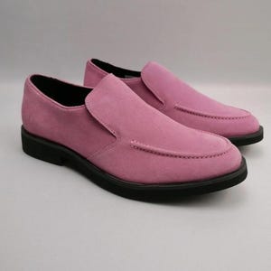 May include: A pair of slip-on loafers crafted from pink suede material. These shoes feature a moccasin-style stitched toe detail, a smooth vamp, and a solid black rubber sole. The interior lining is black, and the overall design is low-profile.