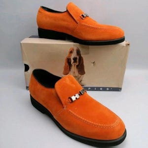 May include: A pair of bright orange suede loafers featuring a silver-toned metal bit detail across the vamp and a black sole. The shoes are displayed with their original cardboard box, which features a graphic of a basset hound and the text PIES.