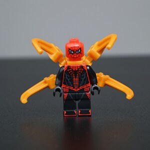 Peut inclure: Une figurine en plastique avec une tête rouge ornée d'un motif de toile d'araignée noire et des yeux blancs. Le torse et les jambes sont noirs avec des détails de toile rouges. Quatre bras mécaniques orange s'étendent à l'arrière.