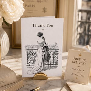 May include: A white greeting card featuring the text Thank You and a black and white illustration of a person in a gown holding a cello on a balcony. The card is displayed in a gold metal stand next to a small sign, a fountain pen, and an envelope.