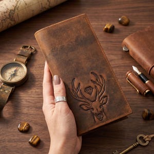 May include: A rectangular brown leather wallet featuring an embossed design of a deer head with a diamond shape between the antlers. The wallet has a smooth, matte texture and is held vertically, showcasing the detailed engraving on the front surface.