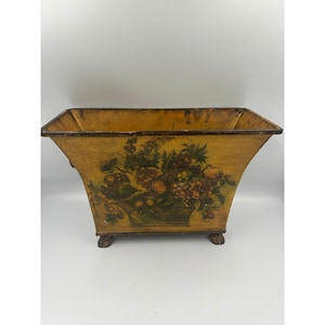 May include: A rectangular metal planter with a flared top and four small feet. The exterior features a distressed mustard yellow finish with a painted central motif of a basket filled with various fruits and green foliage in muted tones.