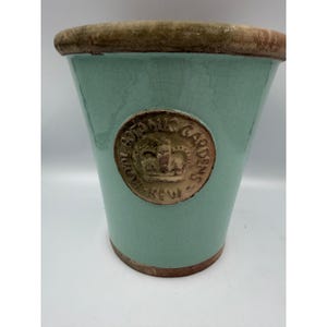 May include: A mint green ceramic plant pot with a crackle glaze finish. The pot features a circular tan medallion on the front embossed with a crown and the text ROYAL BOTANIC GARDENS KEW. It has a wide, unglazed tan rim and a matching base.