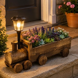 May include: A wooden train-shaped planter with a dark brown stained finish, featuring four circular wheels and a solar-powered lantern attached to the front. The cargo area is filled with a variety of small purple, pink, and green succulent plants.