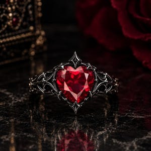 May include: A ring featuring a central heart-shaped red gemstone set within a dark, oxidised silver band. The metalwork displays intricate, pointed filigree details that frame the stone, creating a sharp, ornate appearance with a polished finish.