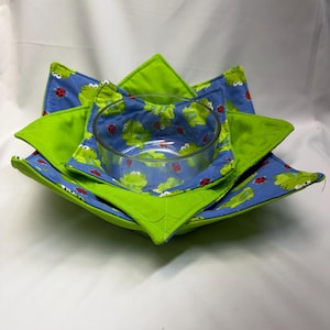 May include: A fabric bowl cosy featuring a blue background with a repeating pattern of green frogs and red ladybugs, accented by solid lime green fabric points. A clear glass bowl is nested inside the quilted, star-shaped fabric holder.