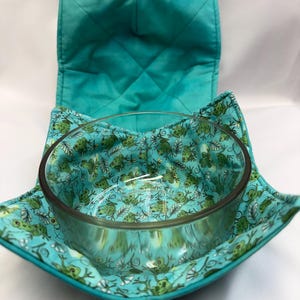 May include: A clear glass bowl sits inside a quilted fabric bowl cosy. The cosy features a vibrant turquoise exterior and an interior lining patterned with small green frogs and white floral accents on a light blue background.