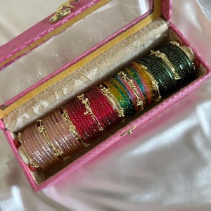 May include: A collection of multi-coloured bangles arranged in a row inside a rectangular pink jewellery box with a clear lid. The bangles feature a textured, glittery finish in shades of brown, red, yellow, green, and dark green with gold accents.