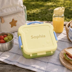 Peut inclure: Une boîte à lunch carrée jaune clair en forme de tranche de pain, avec le prénom Sophia imprimé au centre. Le récipient possède une base verte, des loquets latéraux bleus et une poignée bleue. Il est posé sur un tissu blanc près d'une gourde.