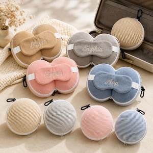 May include: A collection of round, zippered travel cases in beige, gray, pink, and blue, each paired with a matching embroidered sleep mask featuring the names Olivia, Sophia, Emma, or Ava, alongside several striped round cases.