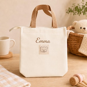Peut inclure: Un sac à déjeuner en toile couleur crème avec deux anses en tissu texturé marron et une fermeture éclair argentée. L'avant présente le prénom Emma brodé en écriture cursive marron au-dessus d'un écusson carré avec un ours dessiné.