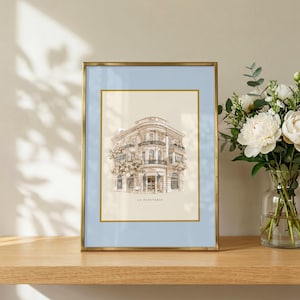 Può includere: Una stampa artistica incorniciata che mostra un'illustrazione dettagliata di un edificio a più piani con balconi e accenti floreali, con la scritta LA PAPETERIE. La stampa è inserita in un passe-partout azzurro e una sottile cornice dorata.