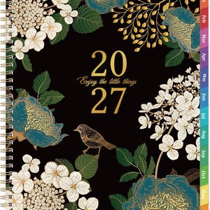 May include: A 2027 spiral-bound planner with a black cover featuring gold text that reads 2027 and Enjoy the little things. The design includes illustrations of white hydrangea flowers, teal and gold peonies, green leaves, and a small brown bird.