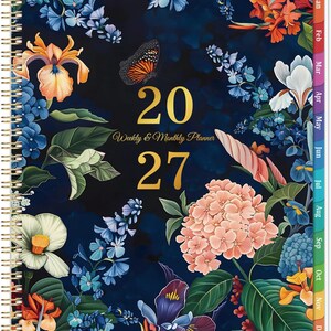 May include: A 2027 weekly and monthly planner with a dark blue background featuring a colorful floral pattern of orange, white, and purple flowers, green leaves, and a butterfly. The cover has gold text and a gold spiral binding on the left side.