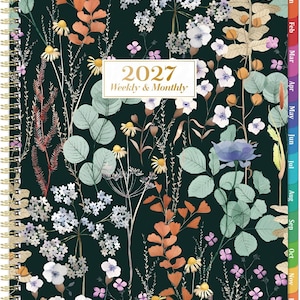May include: A 2027 weekly and monthly planner with a dark green cover featuring a botanical print of various wildflowers, ferns, and leaves in shades of white, blue, and orange. The planner has a gold spiral binding and colored month tabs.