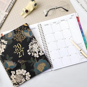 May include: A 2027 spiral-bound planner with a black cover featuring gold and blue floral patterns, a small bird, and the text 2027 Enjoy the little things. The open planner displays a monthly calendar grid with columns for Thursday, Friday, and Saturday.